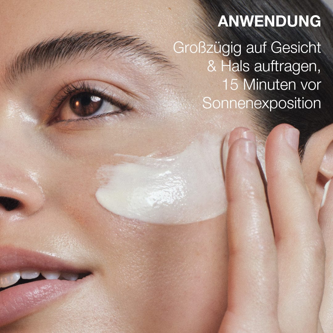 Eine Person trägt Biolumin-C Heat Aging Protector SPF 50 auf ihre Wange auf. Die deutsche Anleitung empfiehlt, es 15 Minuten vor dem Sonnenbad auf Gesicht und Hals aufzutragen.